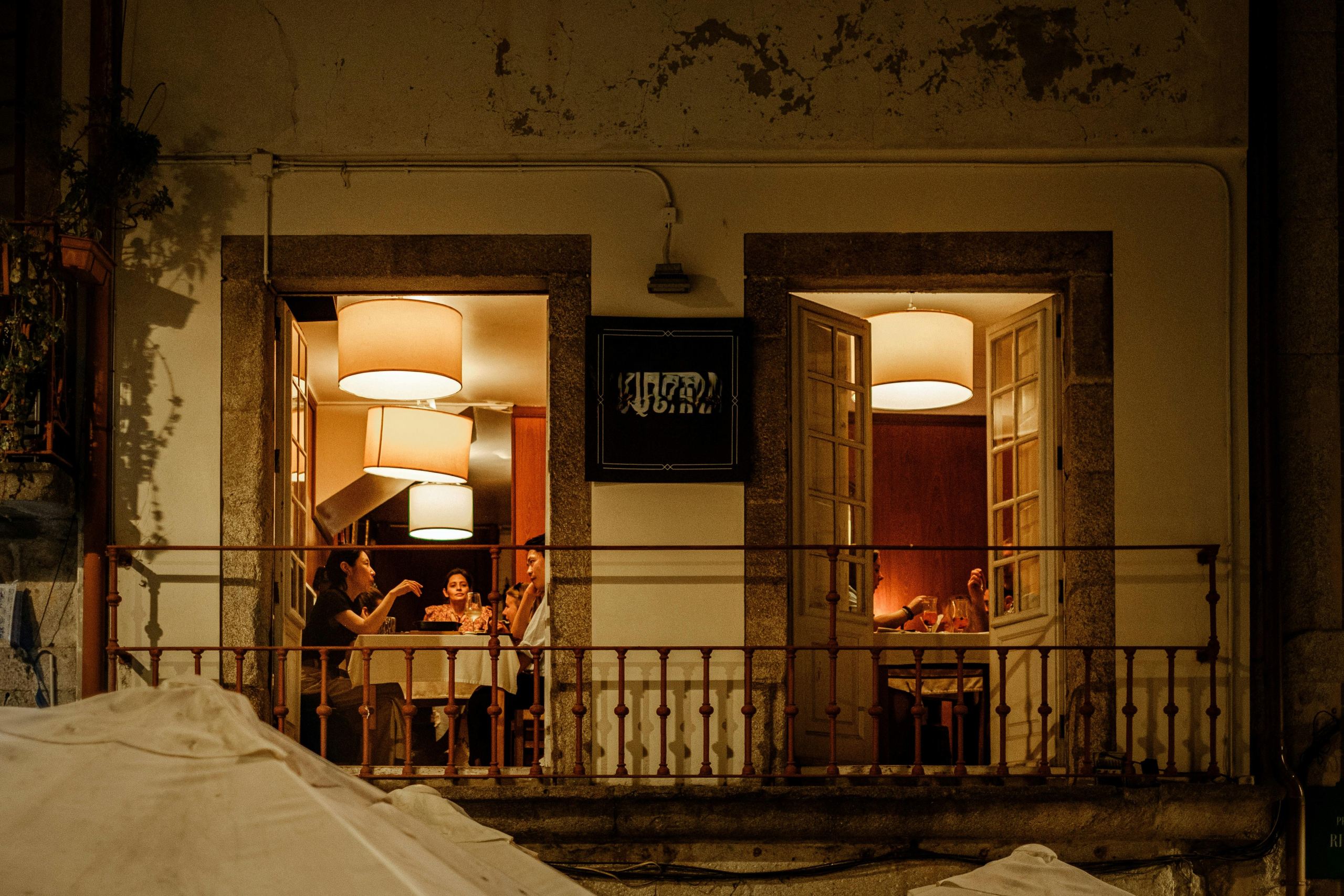 A warm, inviting restaurant scene viewed from outside, featuring diners at candlelit tables, surrounded by soft lighting and open windows.